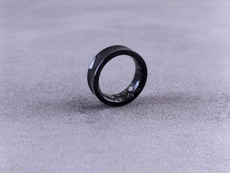 Black titanium smart ring on a textured grey surface - research-derived photograph used to illustrate the form factor; not a personally-owned device.